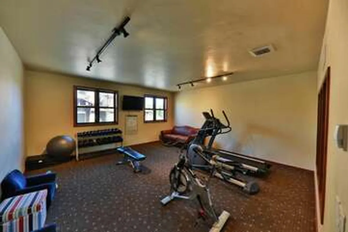 A bright, modern gym area within the clubhouse featuring a range of exercise equipment, weights, a seating area, and large windows letting in natural light. The space is both functional and welcoming, encouraging fitness and well-being in a community setting.