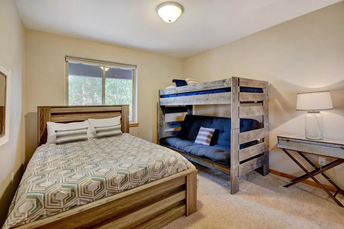 A cozy second bedroom setup with a queen bed and a bunk bed, accompanied by a nightstand, reading lamp, and window. The space is both playful and practical, accommodating families or groups with its flexible sleeping arrangements.