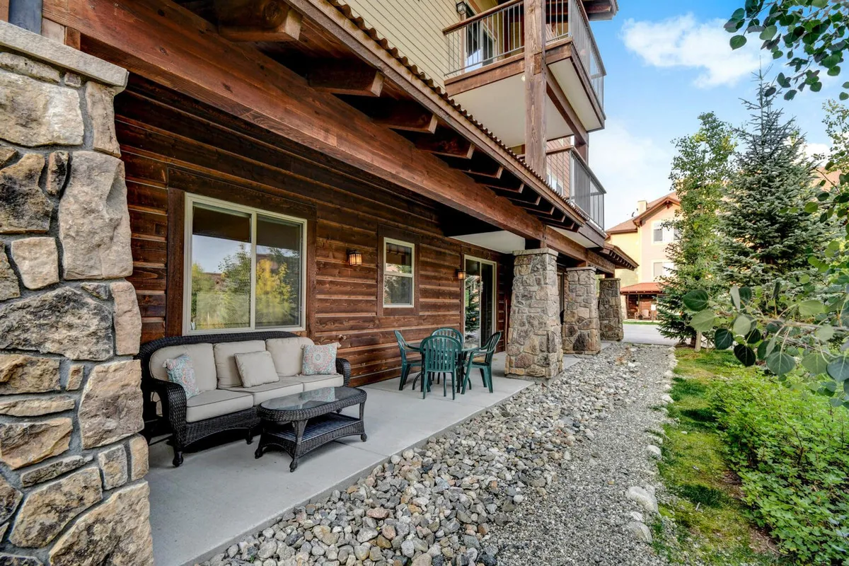 This image shows a private patio area with stone facade, inviting patio seating, green chairs, and a landscaped garden. The patio is directly connected to the condo, surrounded by lush greenery, suggesting a tranquil spot to relax and enjoy the mountain surroundings after a day of adventure.