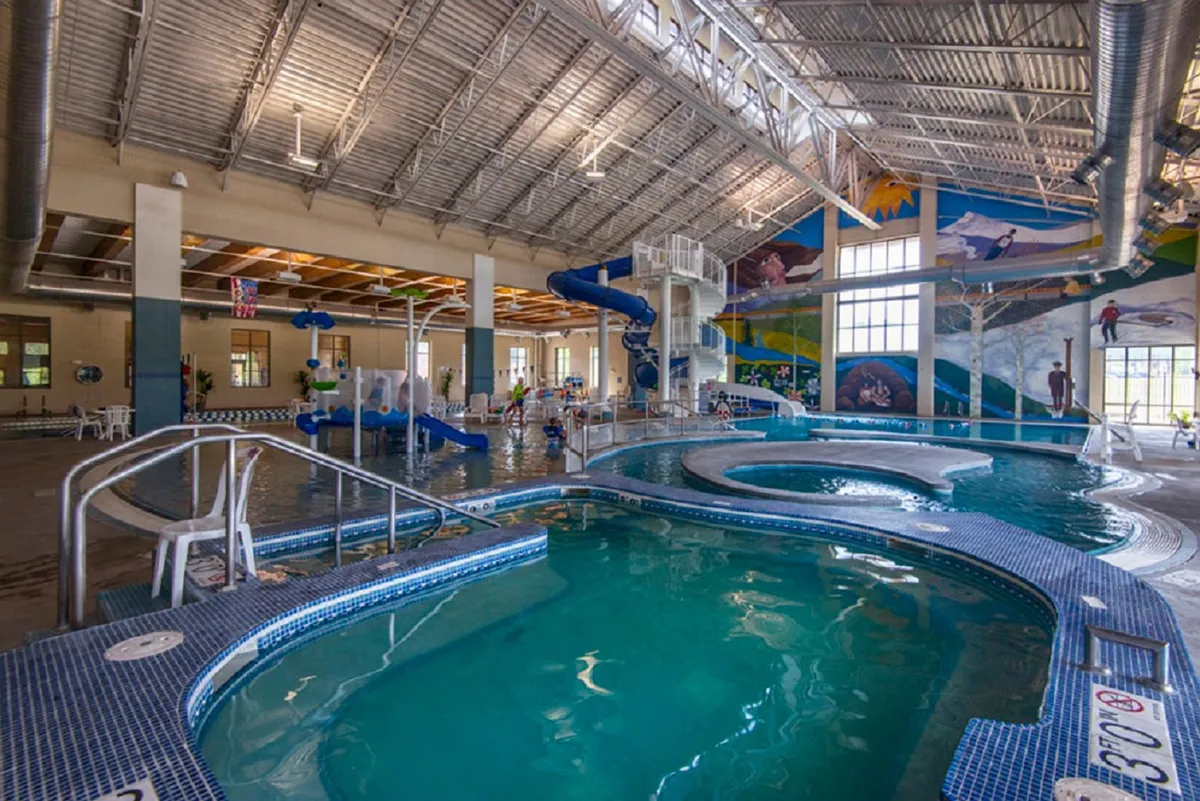An indoor recreation facility featuring bright swimming pools, colorful murals, and water slides, bustling with families and groups enjoying aquatic activities. The space feels joyful and energetic, hinting at opportunities for relaxation and fun breaks during a workday.