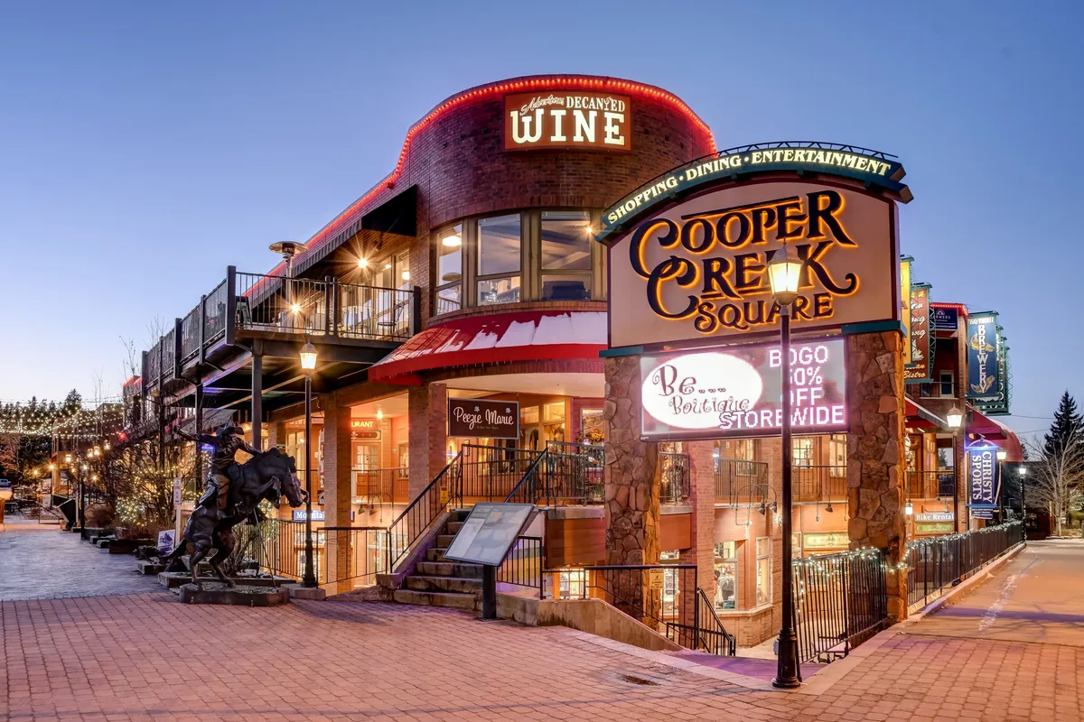 A lively street scene at Cooper Creek Square, featuring a welcoming sign, wine shop entrance, decorative lights, and staircases leading to upper-level shops. The area is bustling with opportunities for shopping, dining, and entertainment.