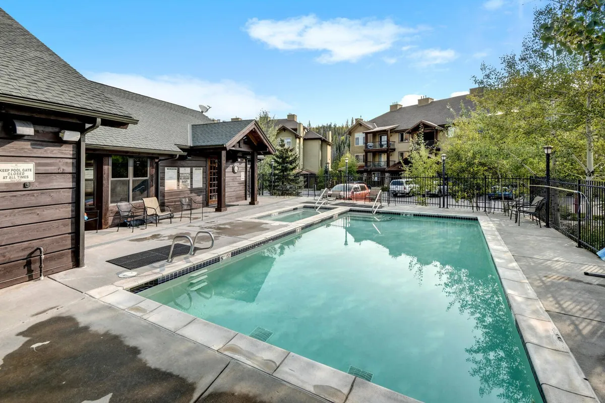 An outdoor heated swimming pool surrounded by lounge chairs and lush greenery, with the clubhouse architecture visible in the background. Residents are seen swimming, sunbathing, and socializing, evoking a relaxed and communal atmosphere ideal for families and friends.