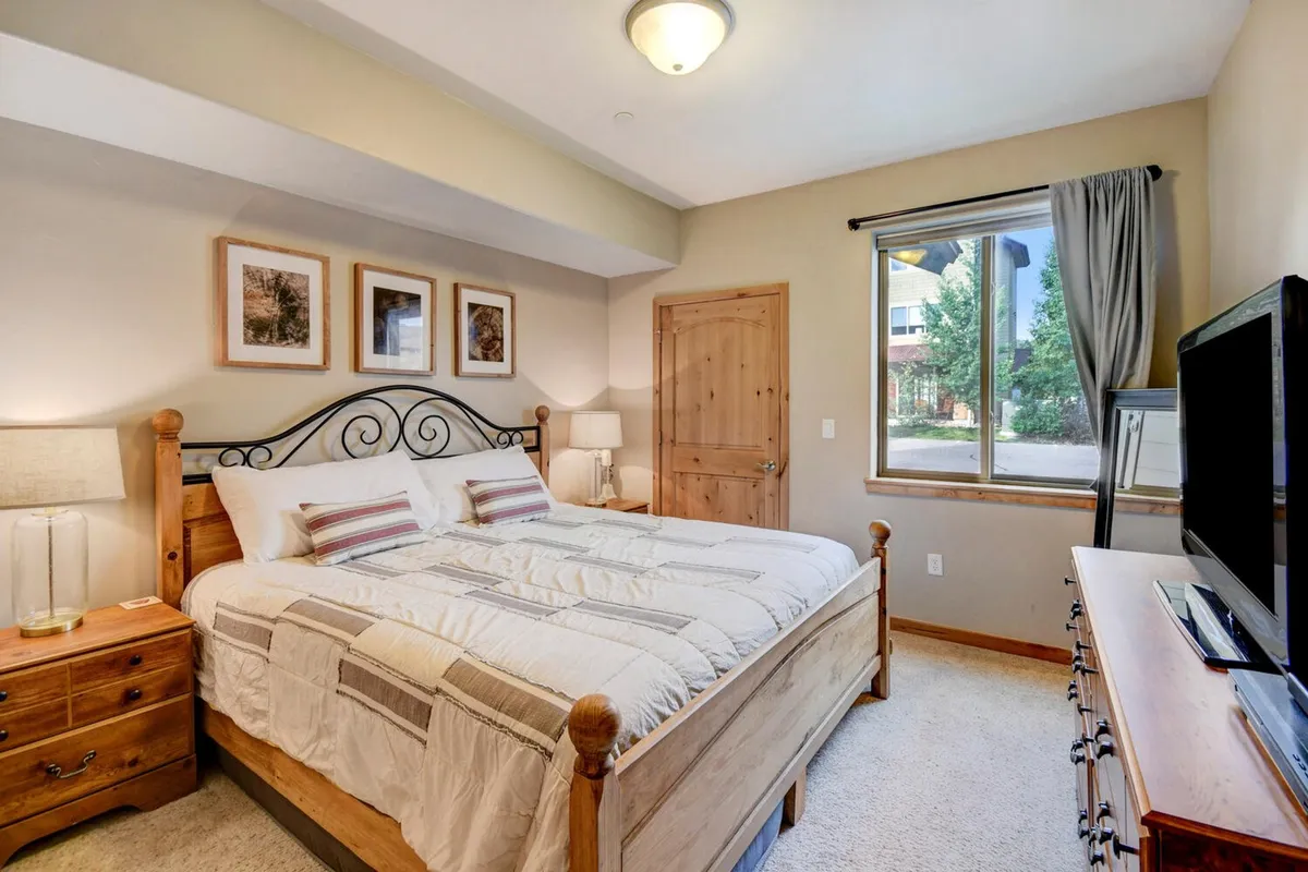 A tranquil bedroom with a neatly made bed, large window with blackout shades, a nightstand, framed artwork, and a wall-mounted television. The soft lighting and tasteful decor create a peaceful and inviting atmosphere.