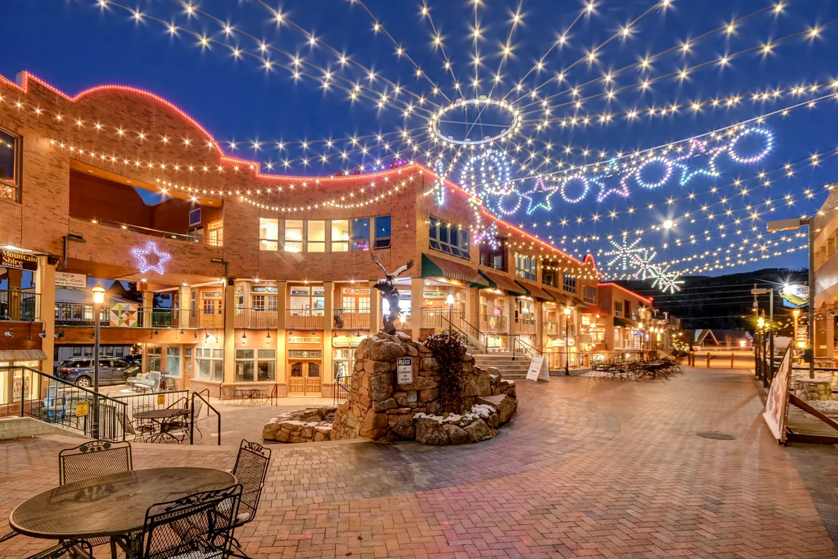 A lively downtown scene at dusk featuring twinkling lights strung above brick pathways, decorative stars, and a central fountain, with people strolling and dining outdoors. The atmosphere is vibrant and welcoming, suggesting easy access to community amenities and a social environment close to the property.