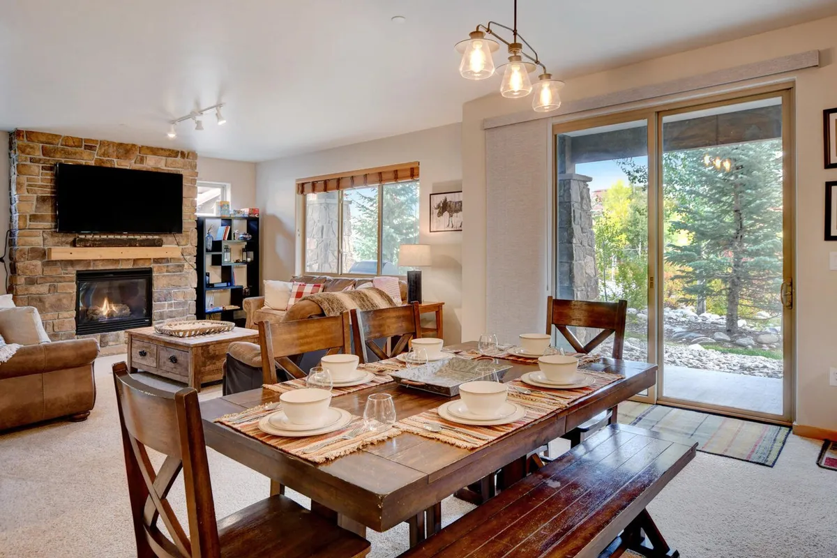 A cozy dining area centered around a dining table with elegant place settings, a striking stone fireplace, and expansive windows looking out onto the trees. The room conveys an atmosphere of warmth ideal for family dinners and intimate gatherings.