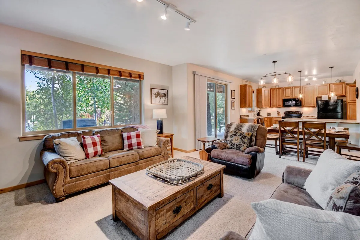 A warm, welcoming open-concept living area featuring comfortable sofas, a wooden coffee table, a kitchen area with bar stools, and large windows showcasing views of nature. The space is set up for group gatherings and relaxation, with seamless flow between the sitting area, kitchen, and dining area.