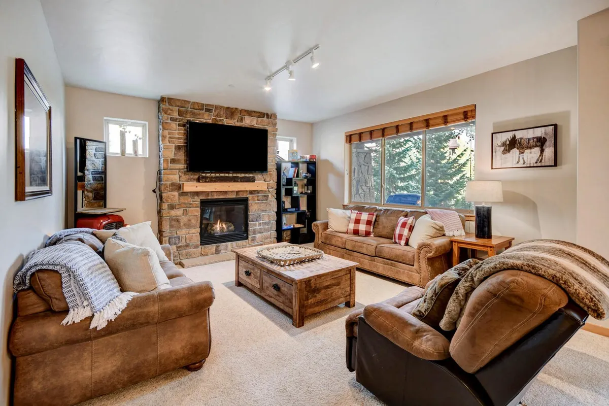 The image showcases a cozy, inviting living room centered around a glowing fireplace, with plush sofas arranged for conversation and relaxation. Large windows frame mountain views, letting in natural light and offering a glimpse of the scenic surroundings. The room is perfect for gathering with family or friends, unwinding by the fire, or enjoying peaceful moments with a good book.